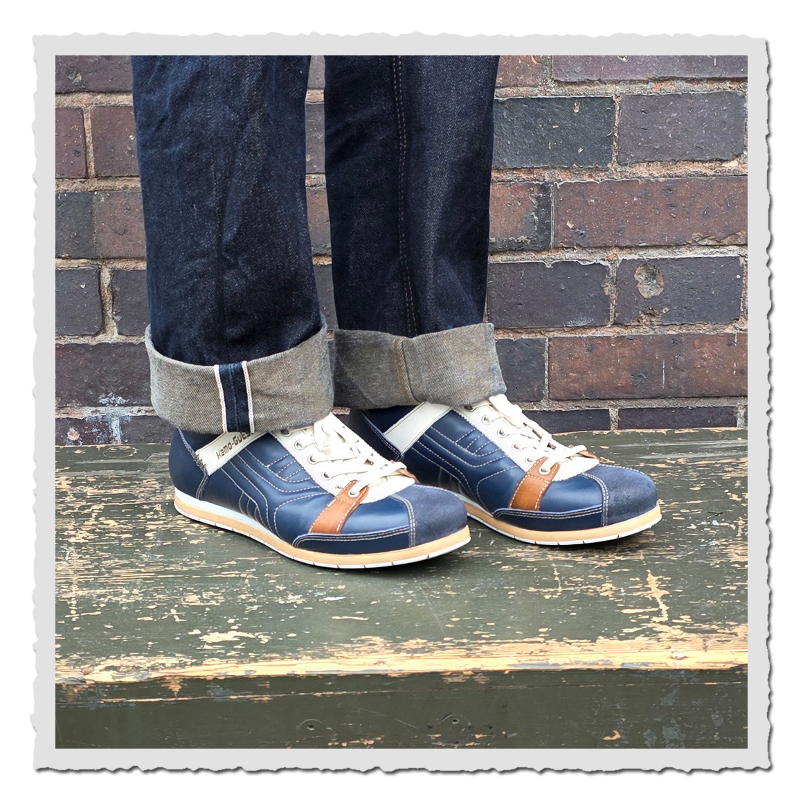 Dunkelblaue Schnürschuhe an einer Person mit umgeschlagenen Jeans, auf grün lackierter Holzfläche vor Backsteinwand.